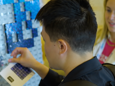 A young man in a black jacket, seen from behind and to the side, holds a small card with a dark blue solar-style tile up to a large mosaic wall of silver and blue pieces. A smiling woman watches in the background.